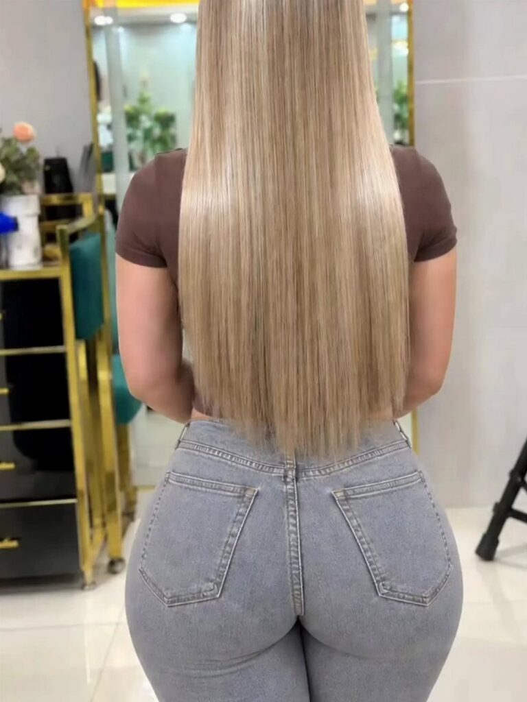 Long straight blonde hair falls to the waist over a brown shirt and gray jeans.