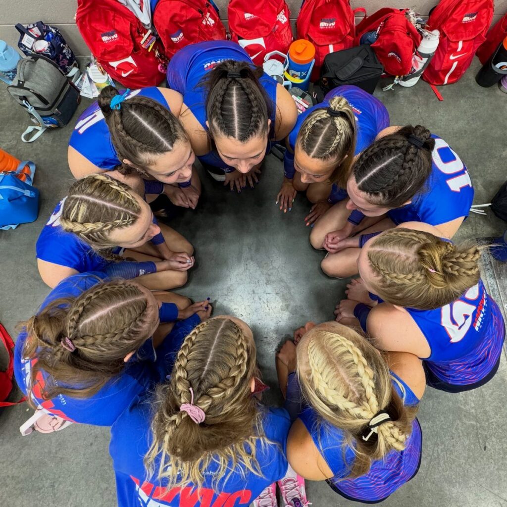 Volleyball players with braided hairstyles kneel together in a team huddle