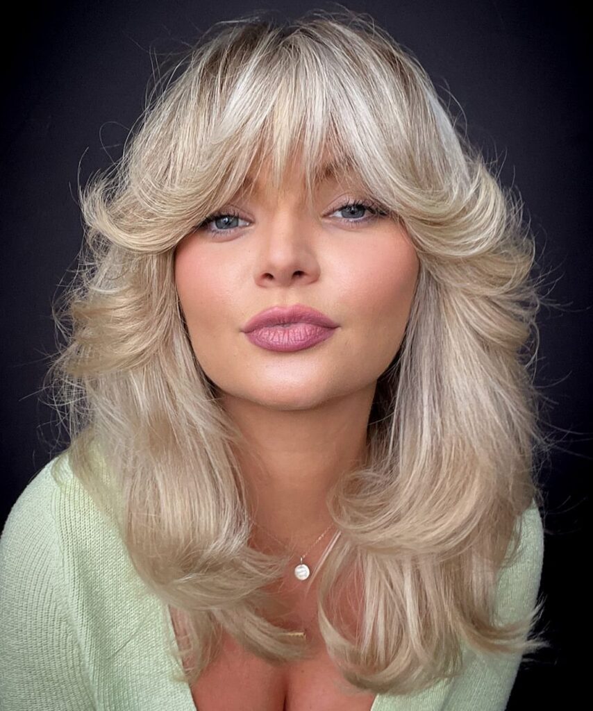 Woman with long blonde hair styled in voluminous rounded layers and sweeping bangs